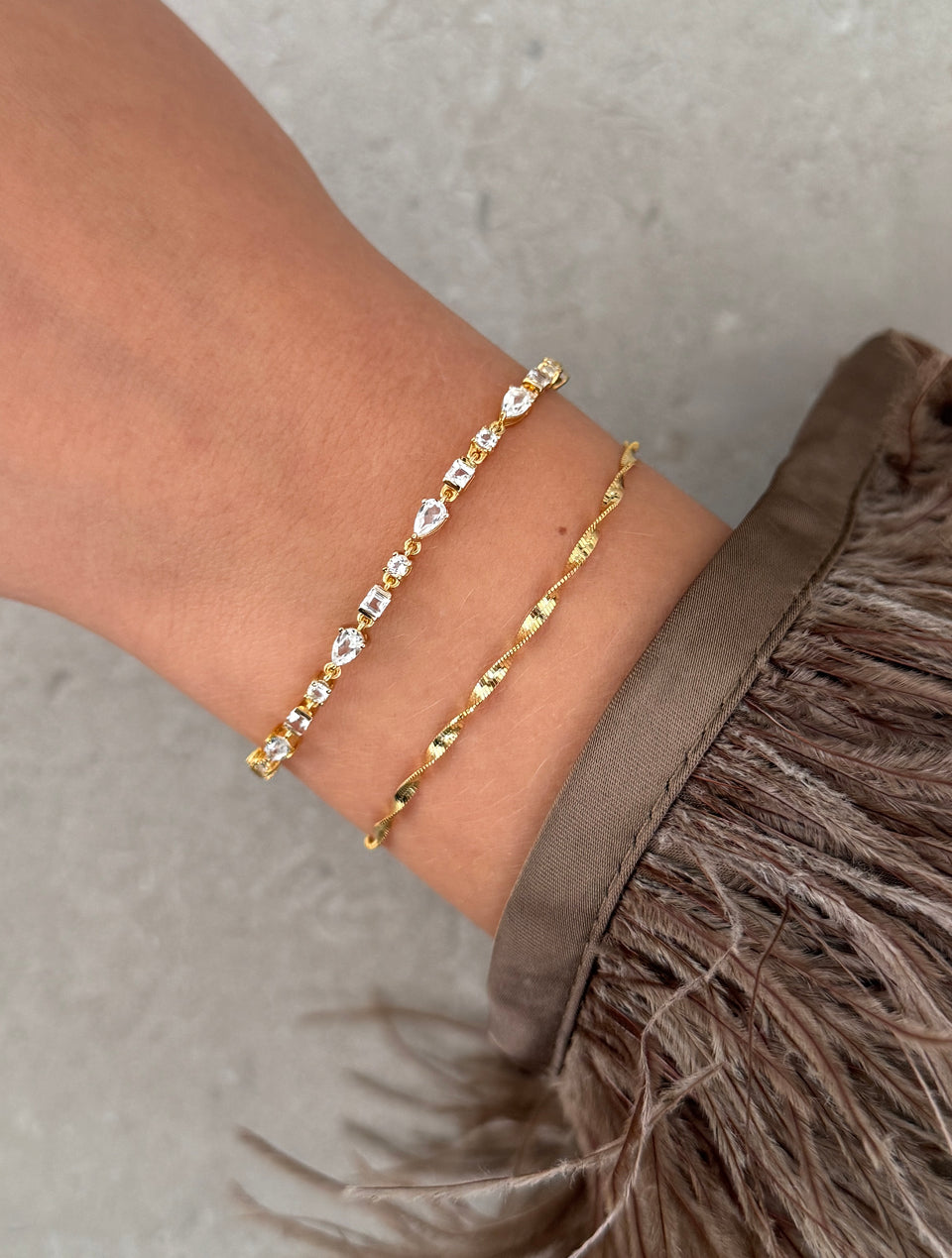A close-up shot of a model's wrist wearing the Herringbone Twisted Bracelet in gold, shown stacked with a delicate gold and clear stone bracelet.