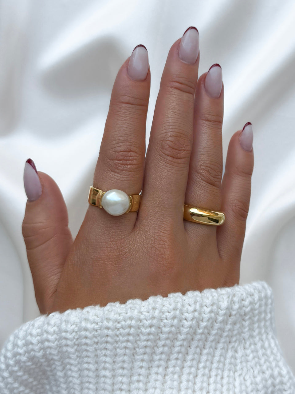 A close-up of a model's hand wearing the Paxton Ring in gold with a large white baroque pearl, alongside a simple bold gold ring.
