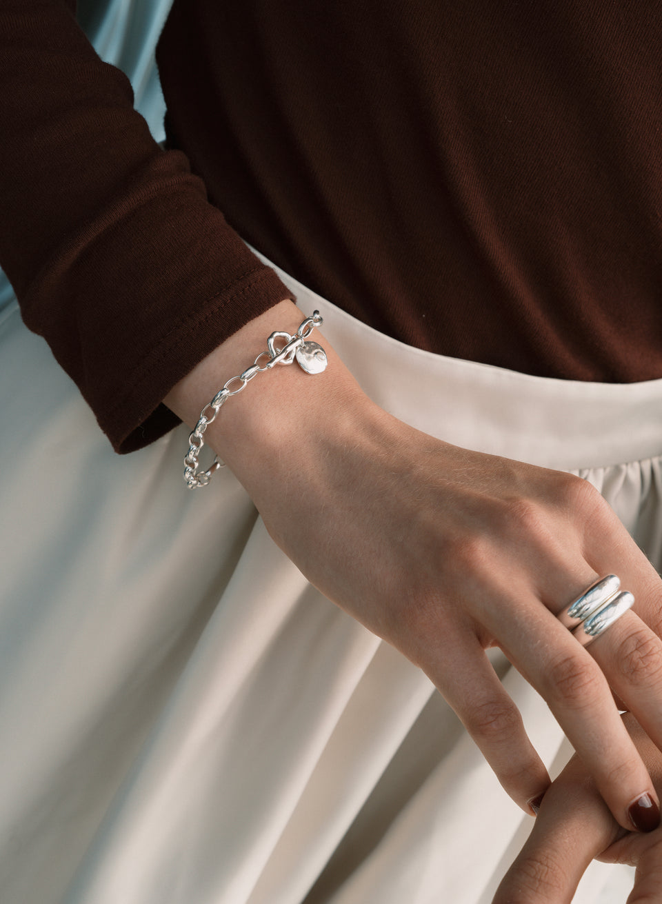 A model wears the Paxton Heavy Bracelet in silver, a chunky link chain with a hammered pendant with a pearl, alongside a wide, double-band silver ring.