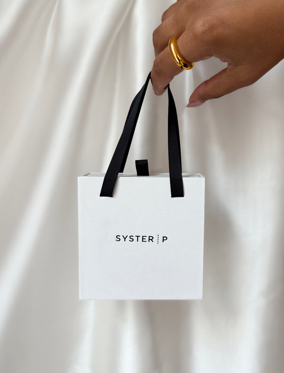 A hand wearing a chunky gold ring holds the white Syster P gift box, showing the beautiful packaging for the Treasure Necklace Set Gold.