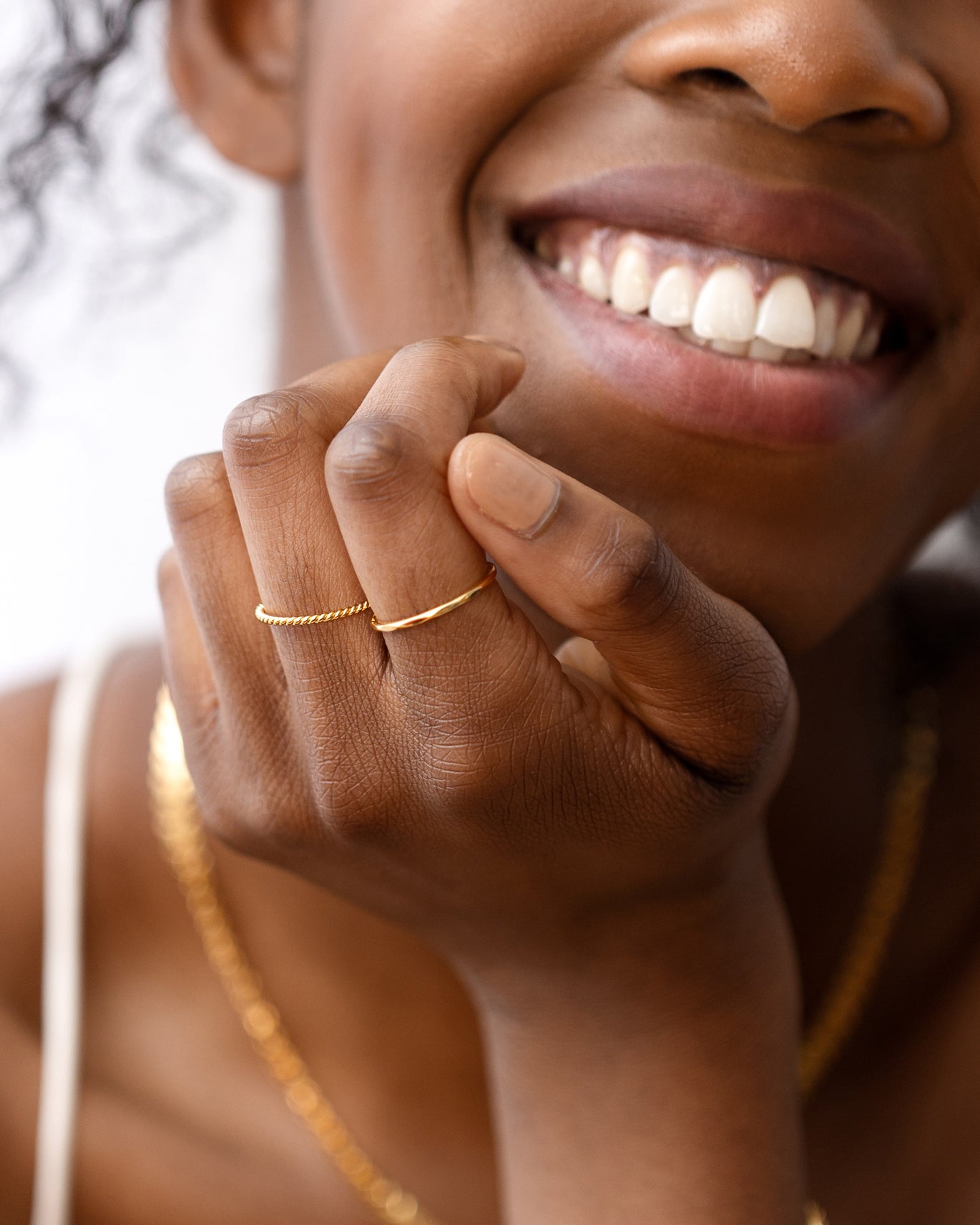 A close-up shot of a smiling model wearing the Tiny Twisted Ring Gold stacked with a simple gold band on one finger, paired with a delicate gold chain necklace.