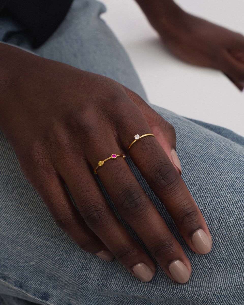 A model wears the gold Treasure Shimmer Ring, featuring a fine band with small, colourful zirconias, styled alongside a second delicate gold ring with a single clear stone.