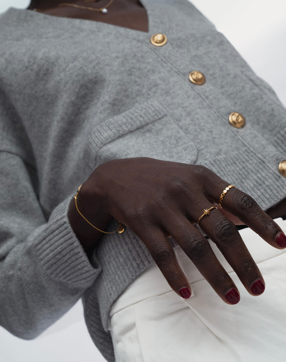 A model styles the gold North Star Ring, which features a delicate star with a central crystal, with a second gold ring that has a dotted design. A fine gold chain bracelet and a delicate pearl necklace complete the elegant look.