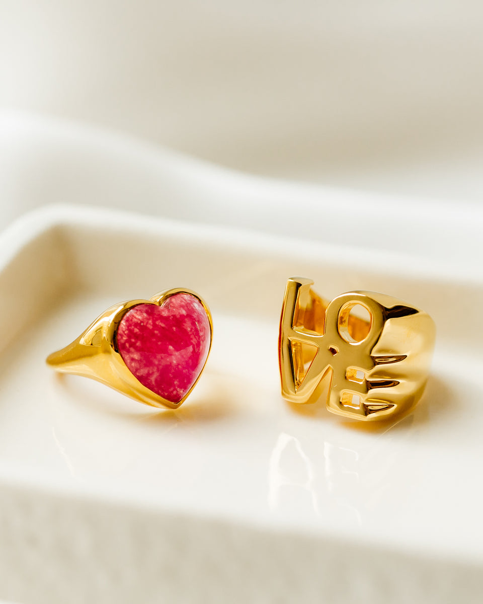 The La La Love Big Heart Ring in gold with its heart-shaped pink quartz stone, styled with the matching 'LOVE' Letter Ring in gold.
