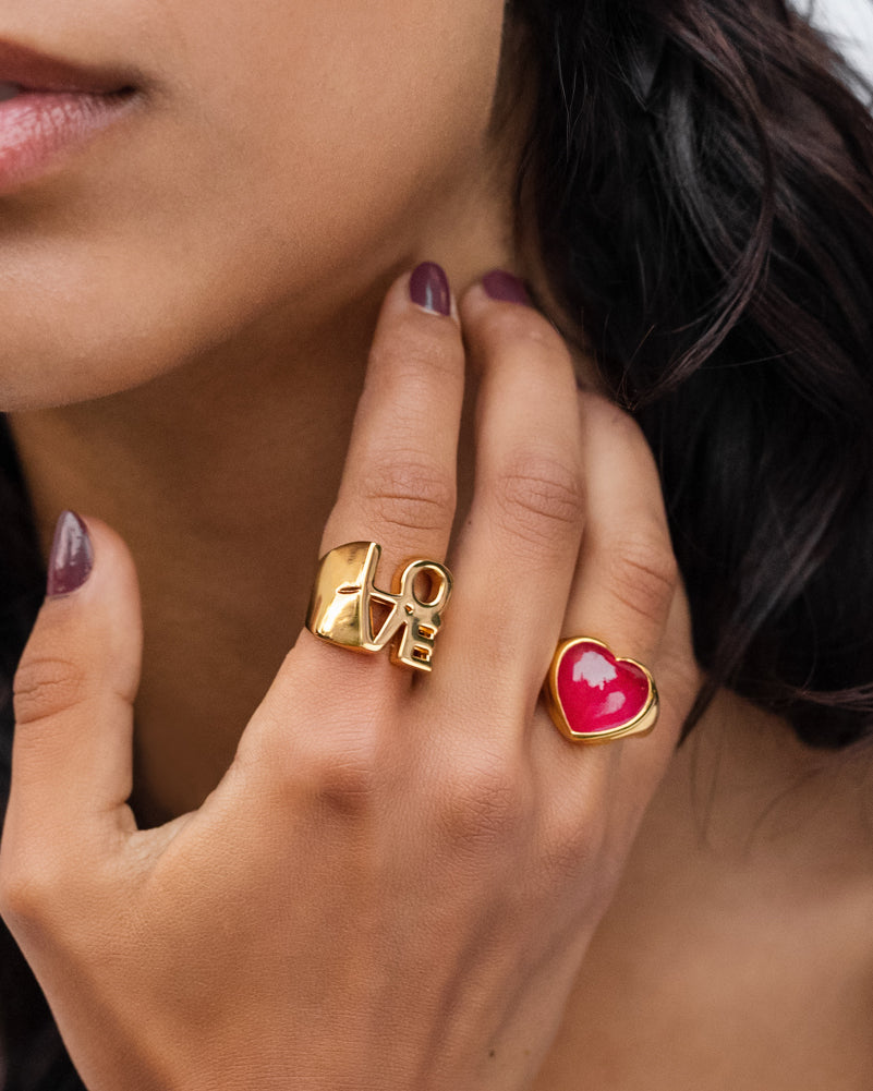 A model's hand showcasing the gold La La Love Big Heart Ring with a bright pink quartz heart, styled alongside a chunky gold 'LOVE' ring.
