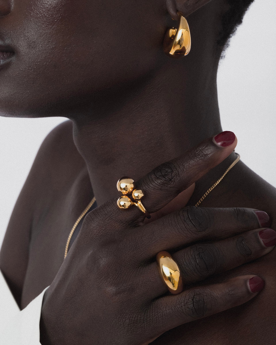 A close-up of a model wearing the Universe Ring Gold, which features three spheres, on her index finger. She is also styled with a chunky gold band, a thick gold hoop earring, and a delicate gold chain necklace.