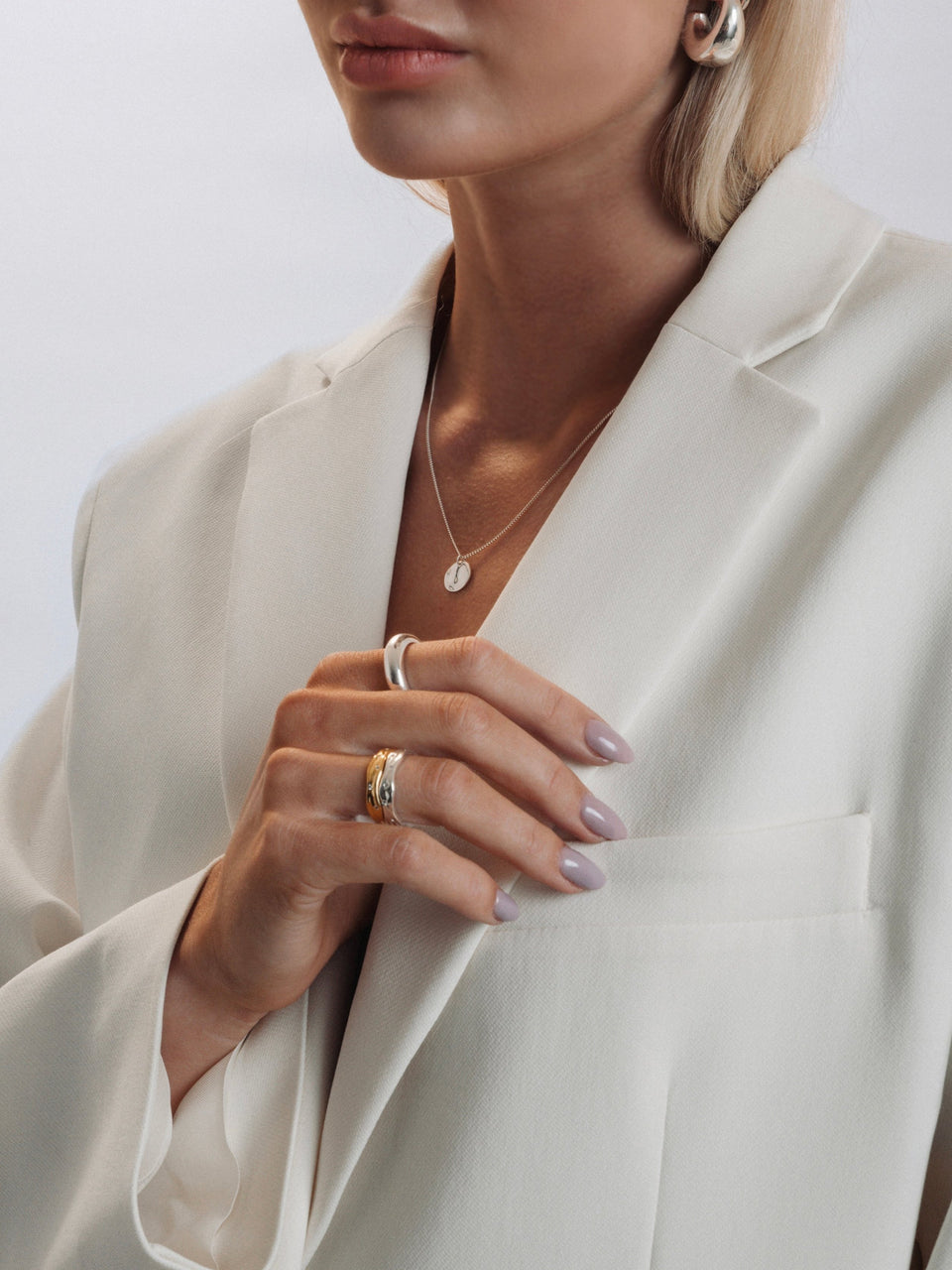 A model styles a mix of Syster P jewelry with a white blazer, featuring the Melting Narrow Ring in silver stacked with the gold version and a plain band. The look is completed with a wide silver ring on another finger, a delicate silver pendant necklace, and chunky silver hoop earrings.