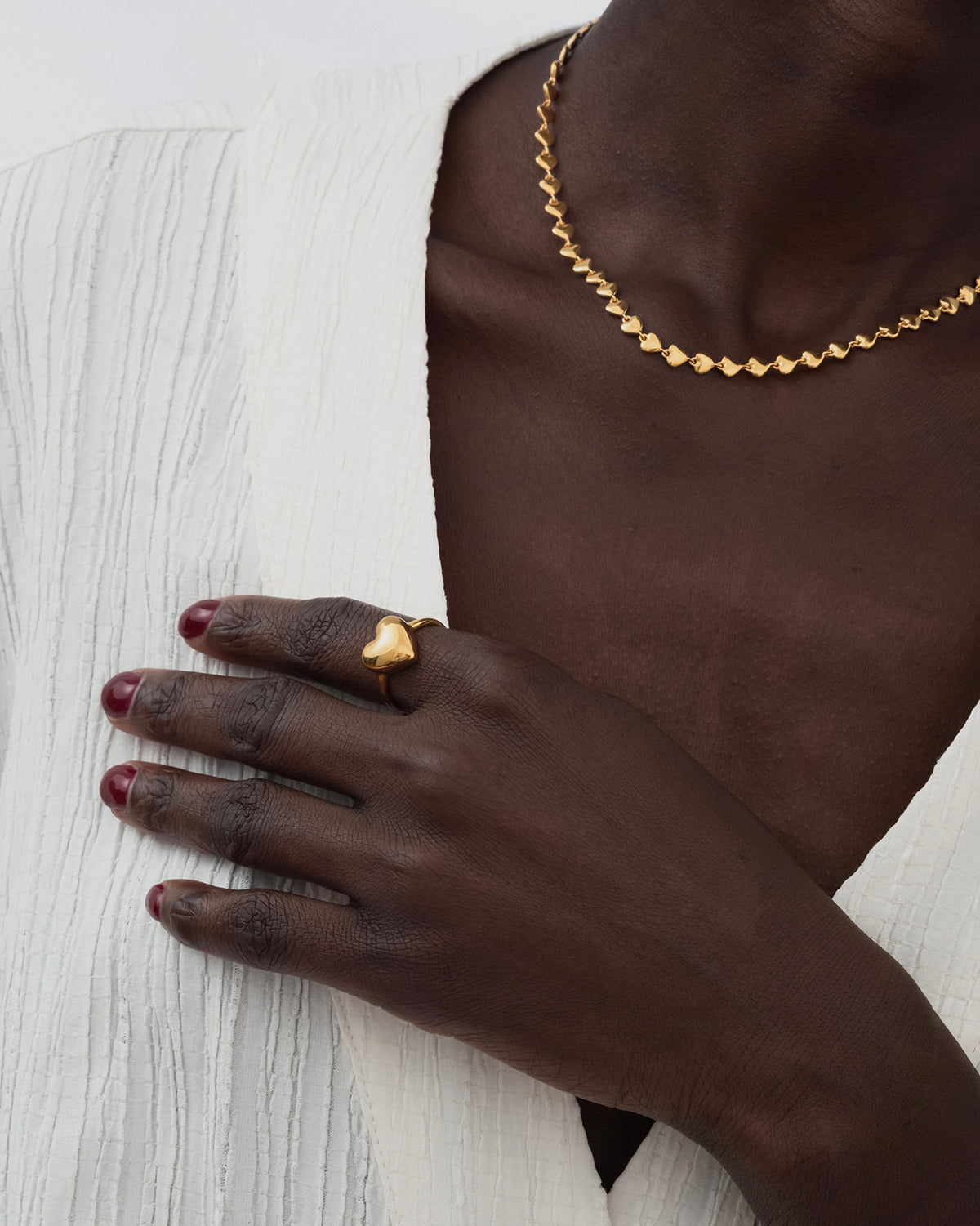 A close-up of a model wearing the Darling Ring in gold, a statement piece with a large heart, paired with a delicate gold necklace made of small, linked hearts.