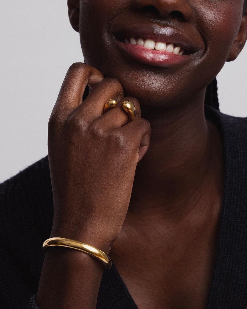 A close-up shot of a model wearing the Bolded Bangle in gold on her wrist, paired with a matching open-ended gold ring with drop-shaped tips on her finger.