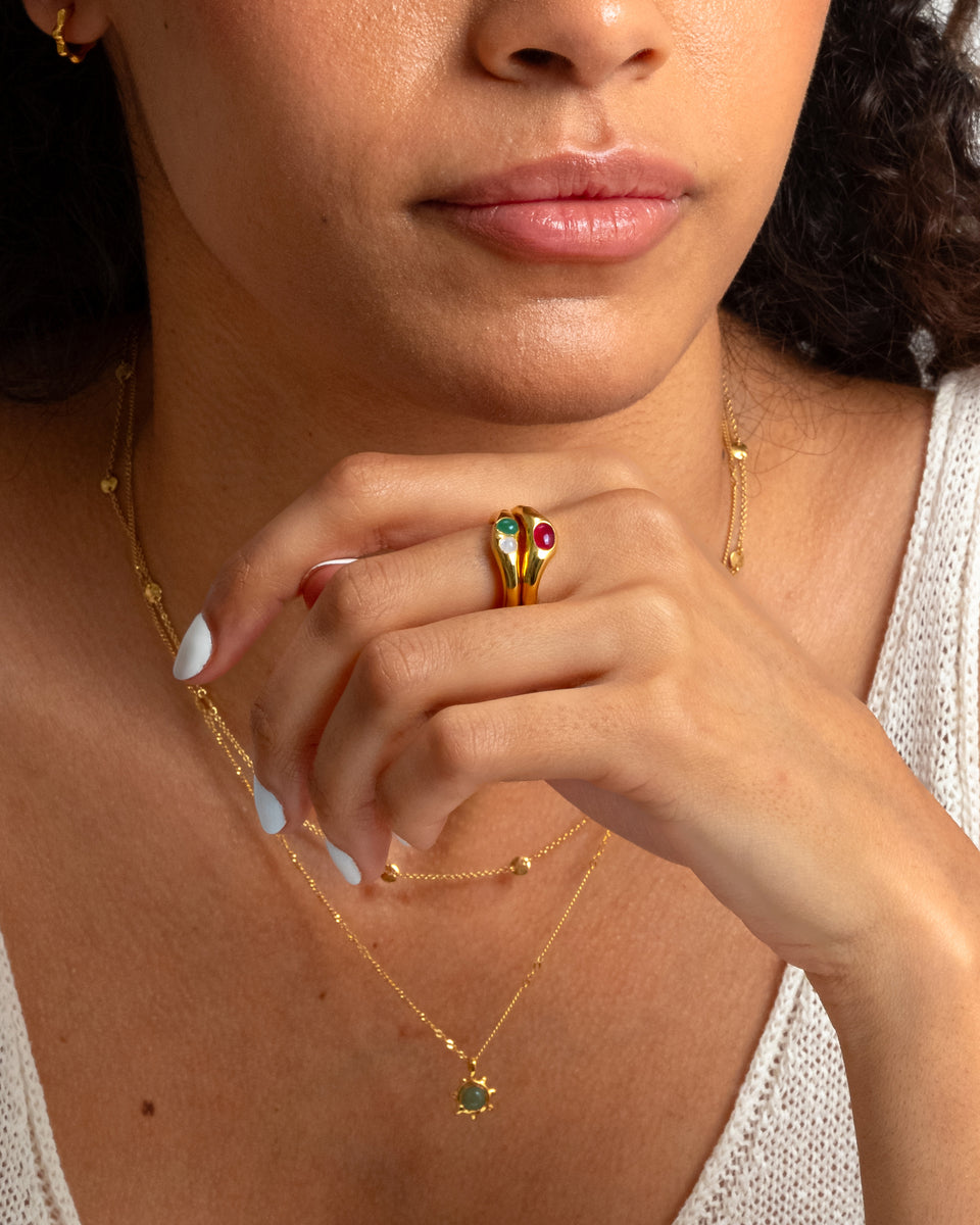 A close-up of a model wearing two stacked Florentina Rings in gold, one with green jade and moonstone and the other with a red stone, along with layered gold necklaces and a small gold hoop earring.