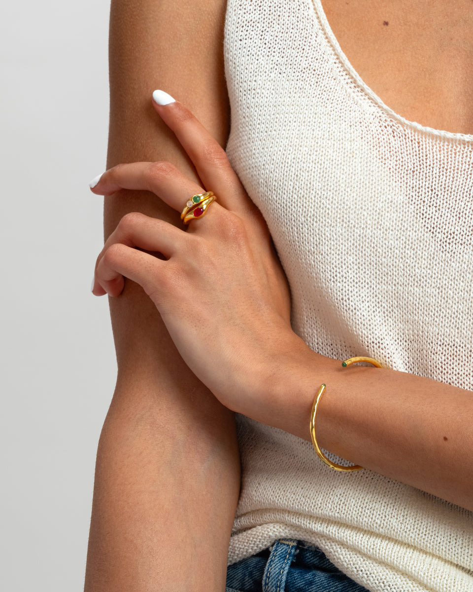 A shot of a model wearing two stacked Florentina Rings in gold, one set with a green jade and a moonstone and the other with a red stone, along with a gold cuff bracelet.