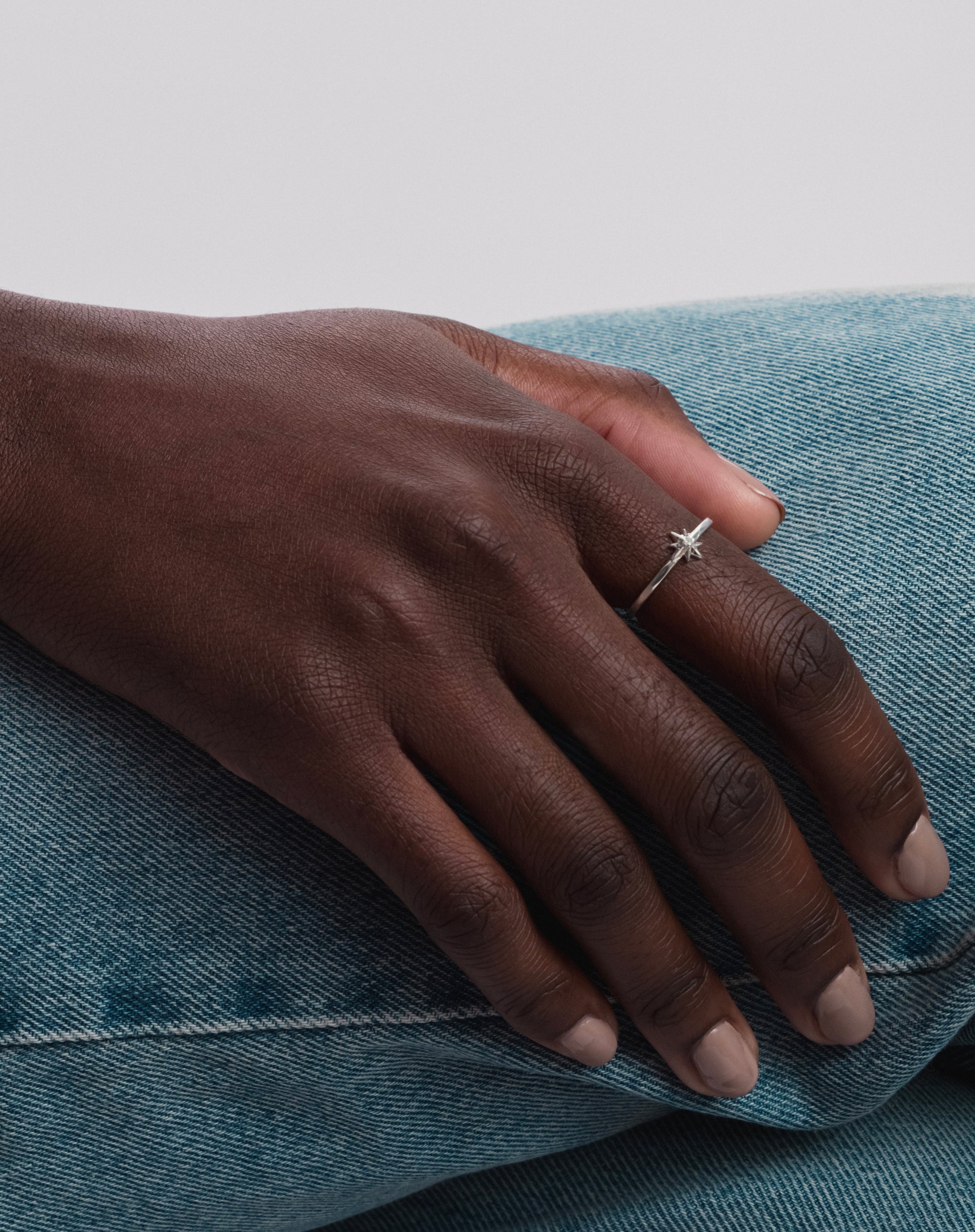A model wears the North Star Ring in silver, a delicate piece with a star and a central crystal, shown on her finger as her hand rests on blue denim jeans.