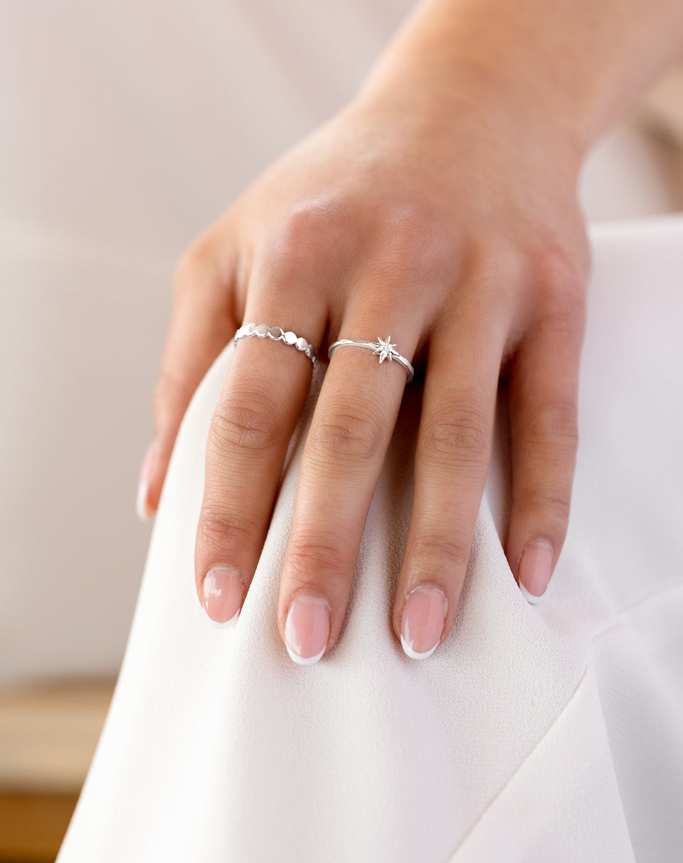 A close-up shot of a model's hand wearing the North Star Ring in silver on the middle finger, featuring a small star with a crystal, alongside another silver ring with a pattern of flat circles on the index finger.