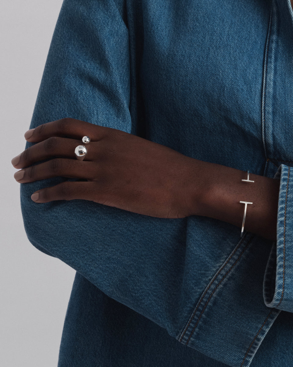 A model wears the Bolded Drops Ring in silver, featuring two polished drop-shaped orbs, styled with a slim silver cuff bracelet with T-bar ends.