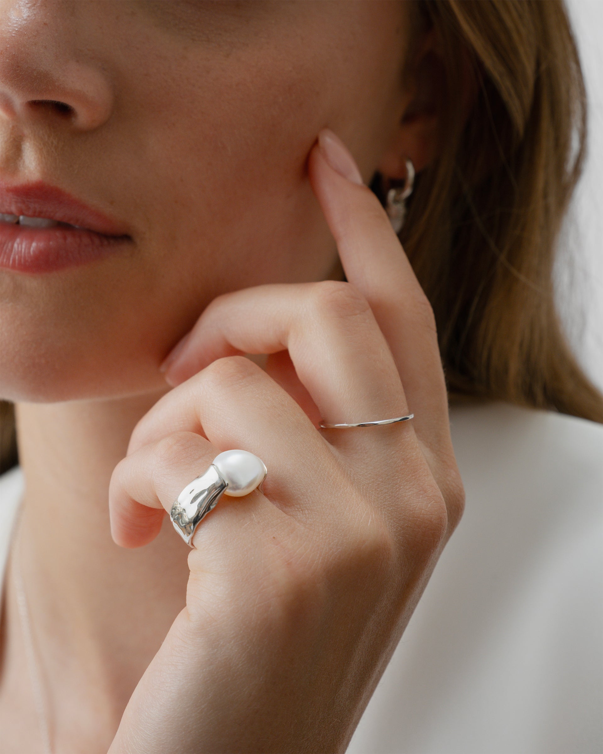 A model wears the Tiny Ultrathin Ring Silver, a simple band, on her index finger, alongside a bold, sculptural silver ring with a large pearl on her ring finger, and a small silver hoop earring.