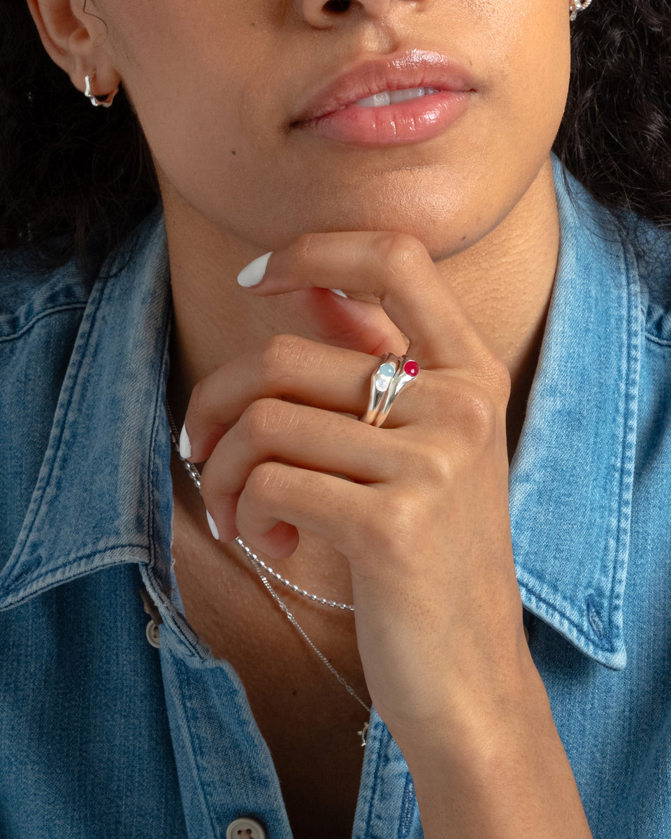 A close-up of a model wearing the Florentina Ring in silver, with its aquamarine and moonstone gems, stacked with a matching silver ring featuring a red stone. She also wears a fine silver chain necklace and small silver hoop earrings.