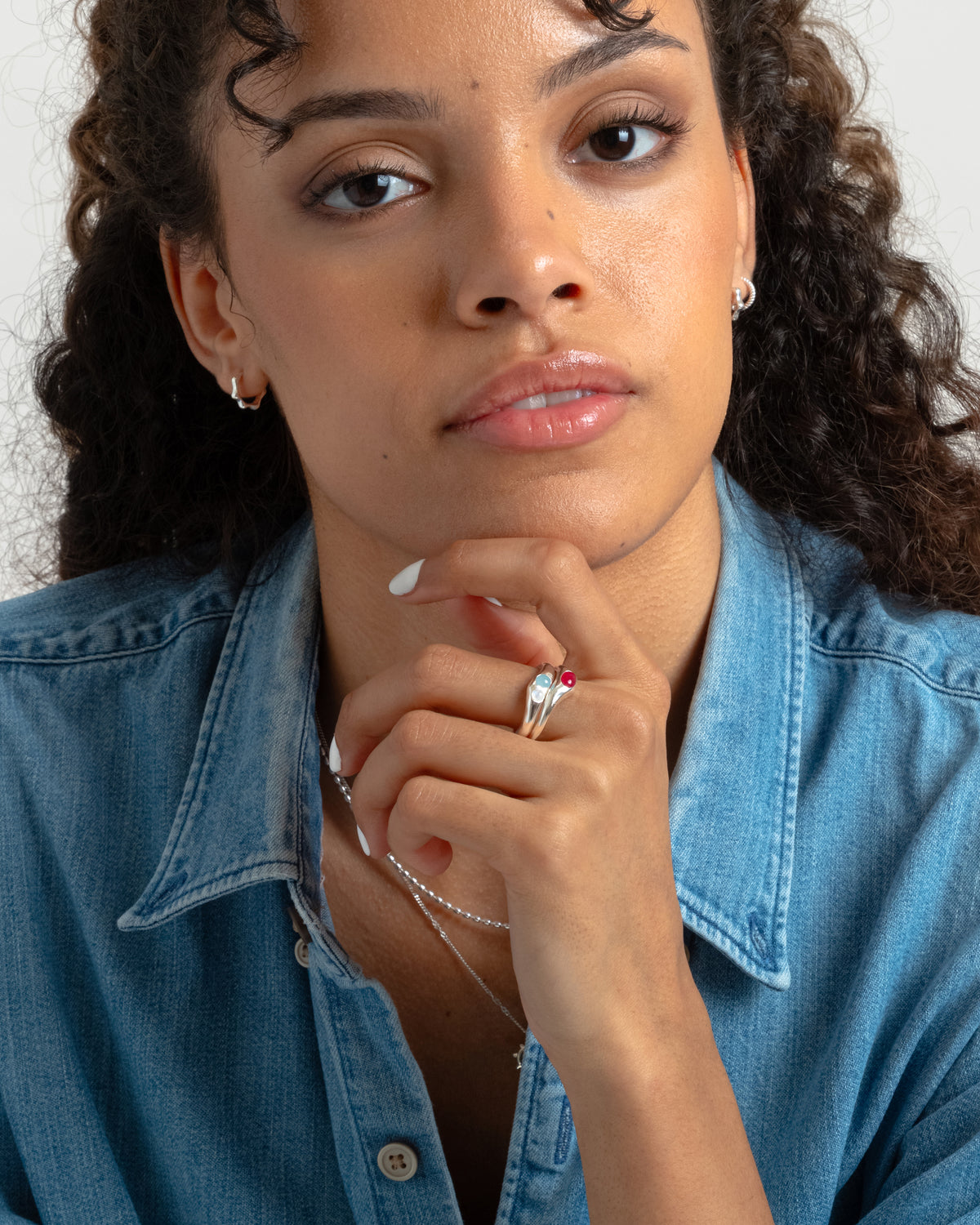 A model poses with her hand to her chin, showcasing a stack of three silver Florentina rings with pink quartz, aquamarine, and moonstone gems, complemented by a silver ball chain necklace and small silver hoops.
