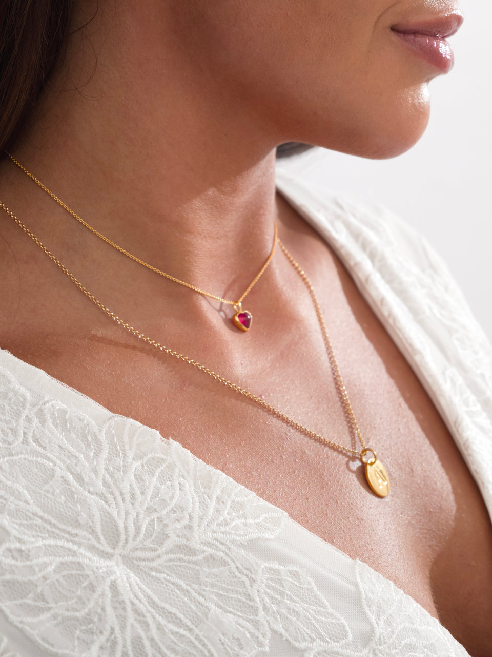 A close-up of a model wearing two layered gold Syster P necklaces. The shorter one features a pink heart pendant, and the longer one is a Beloved Rolo Chain with a gold letter pendant.