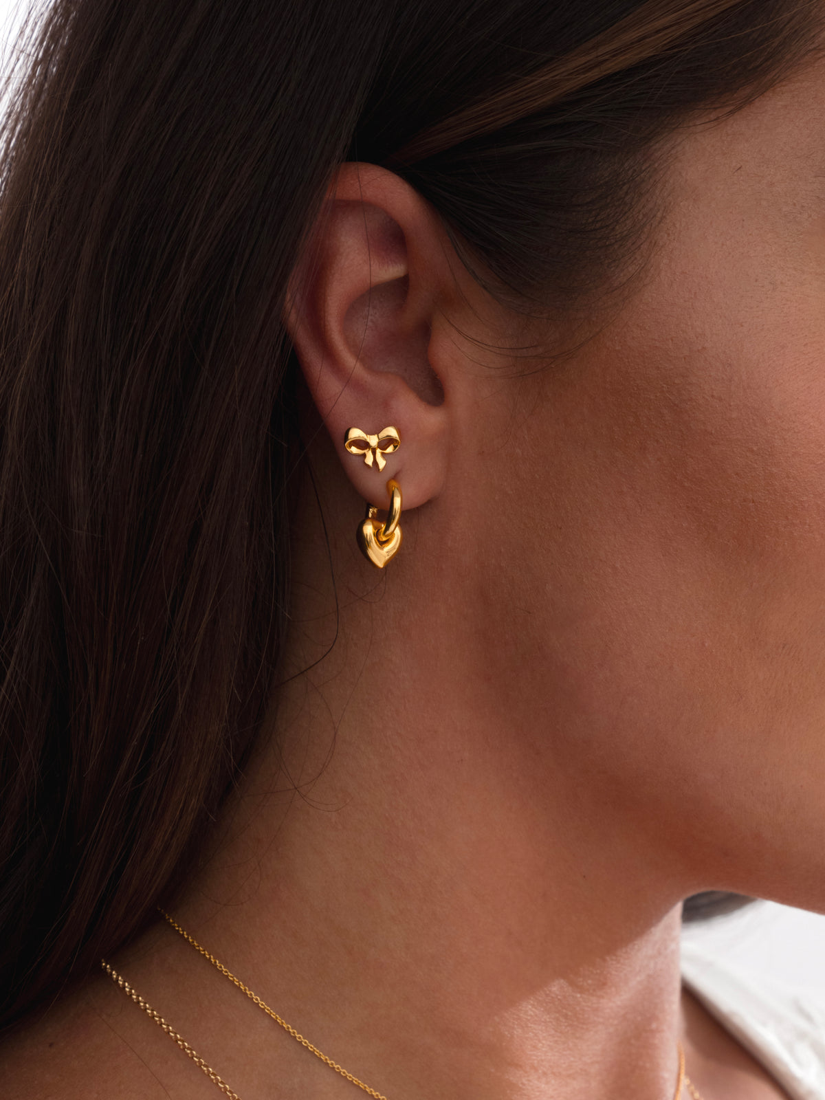A close-up on a model wearing a gold bow stud earring and the La La Love Fat Heart Earrings, complemented by a layered set of fine gold necklaces.