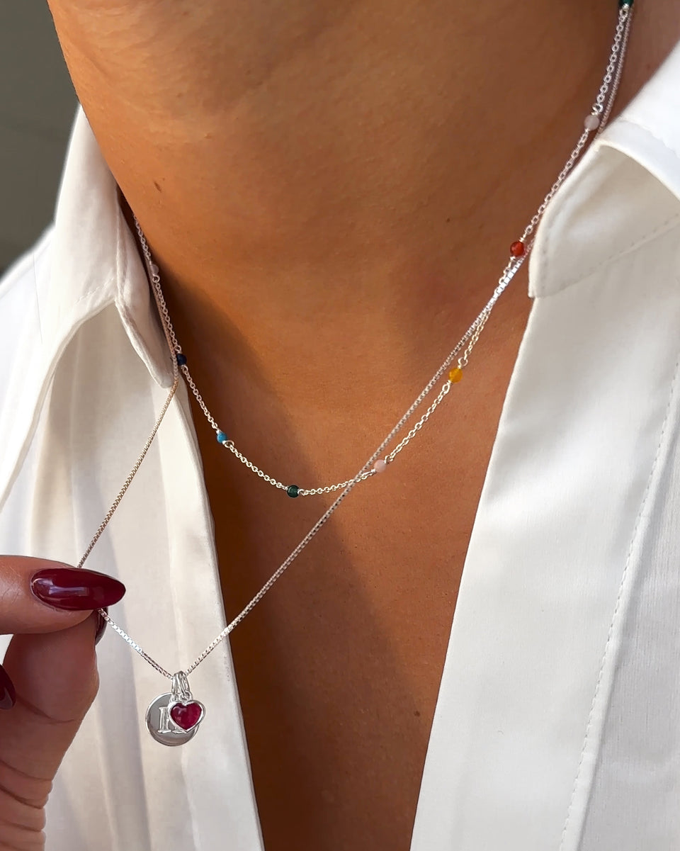 A close-up on a model wearing the Santa Monica Necklace with colorful gemstones, layered with a silver box chain necklace featuring a mini letter 'L' charm and a pink quartz heart charm.