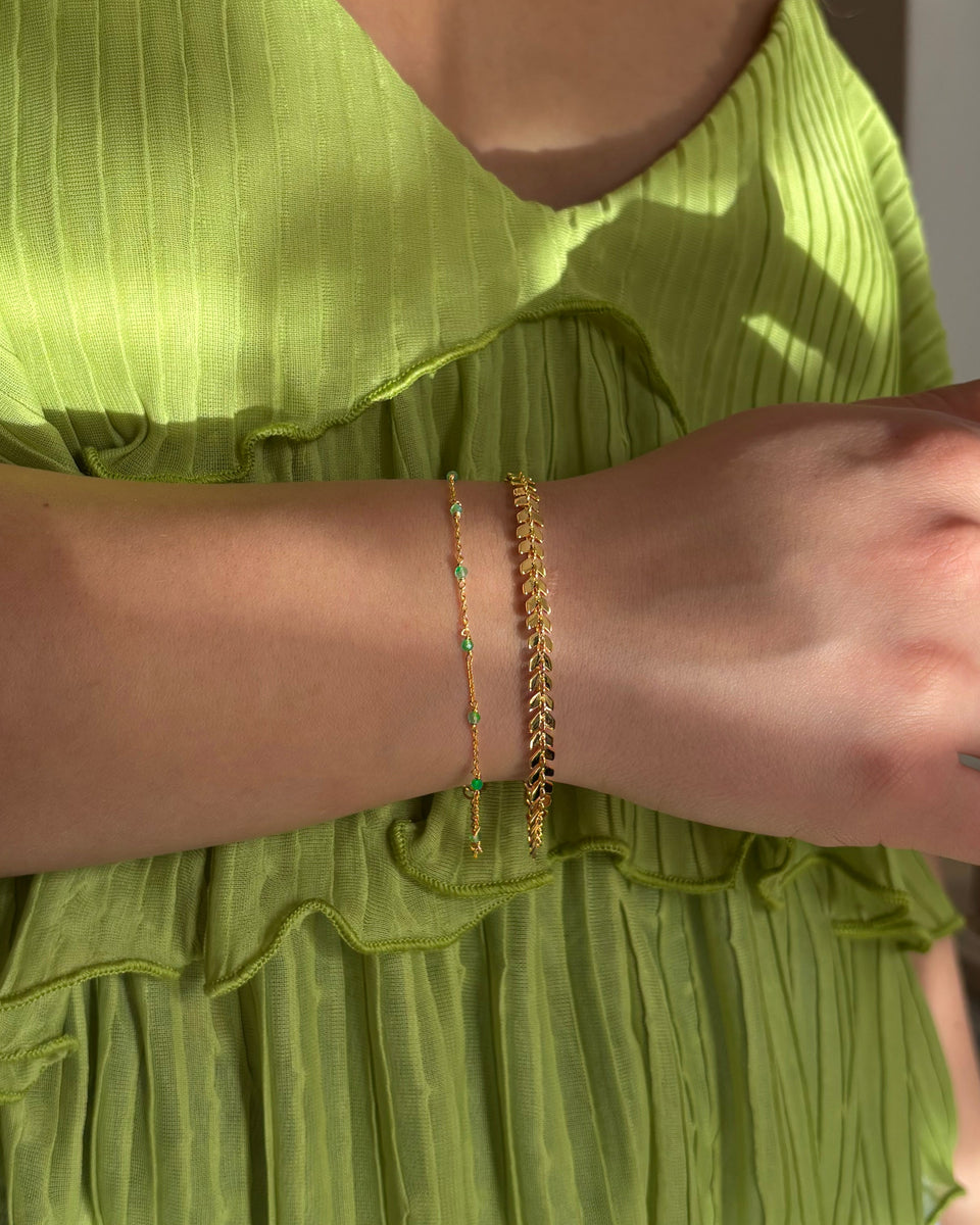 A model's wrist showcasing a layered look with two gold bracelets: the delicate Santa Monica Bracelet with green onyx stones and the Olivia Bracelet featuring a leaf-like pattern.