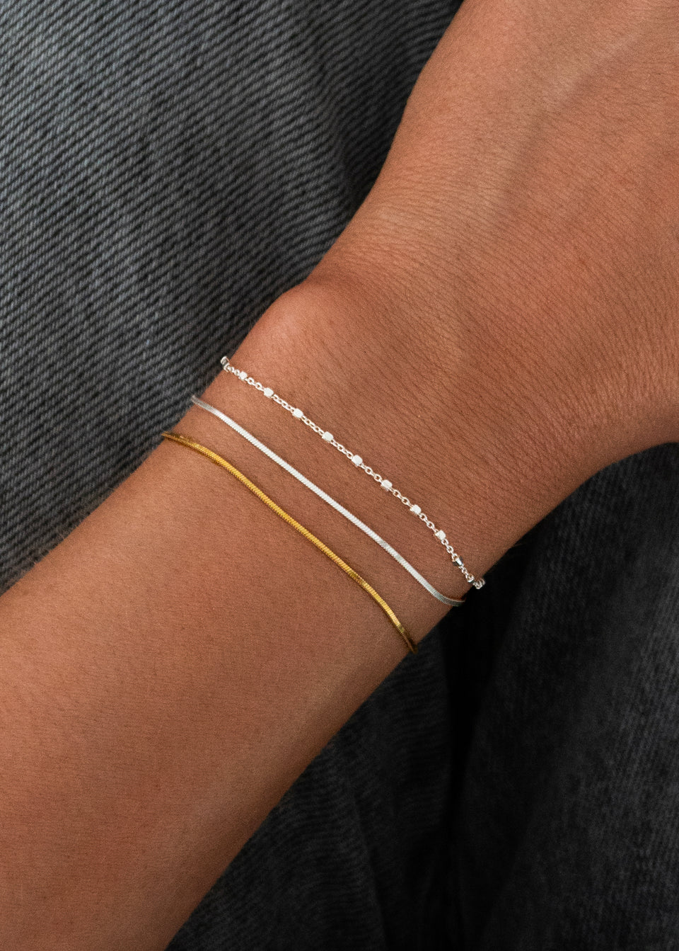 A close-up of a model's wrist wearing a stack of three delicate chain bracelets: the Sheer Station Bracelet in silver, the Sheer Mirror Bracelet in silver, and the Sheer Mirror Bracelet in gold.