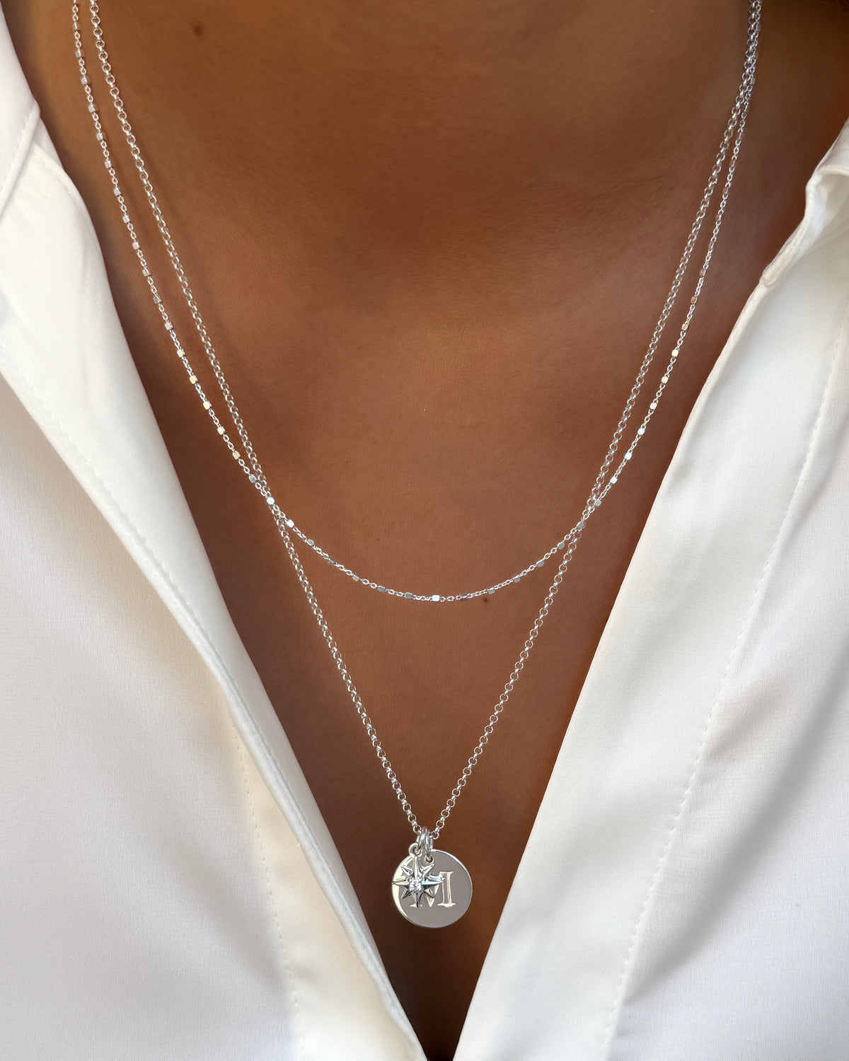 A close-up of a model wearing two layered silver necklaces. The shorter necklace is a delicate station chain, and the longer rolo chain is adorned with a round letter 'I' pendant and a compass star pendant.