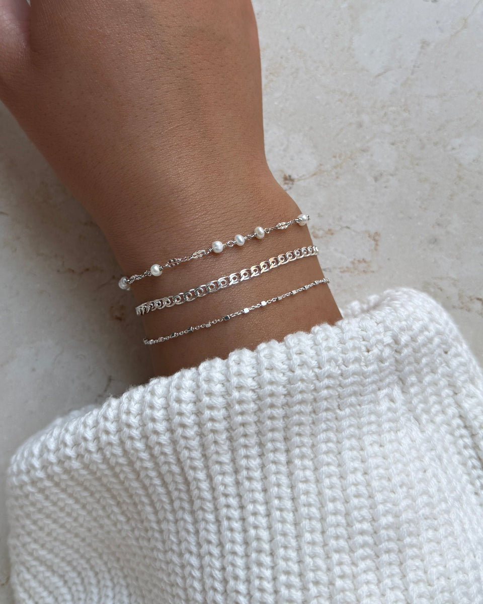 A close-up of a model's wrist wearing a stack of three silver bracelets: the Aurelia Bracelet with pearls, the Sheer Love curb chain bracelet, and the delicate Sheer Station bracelet.