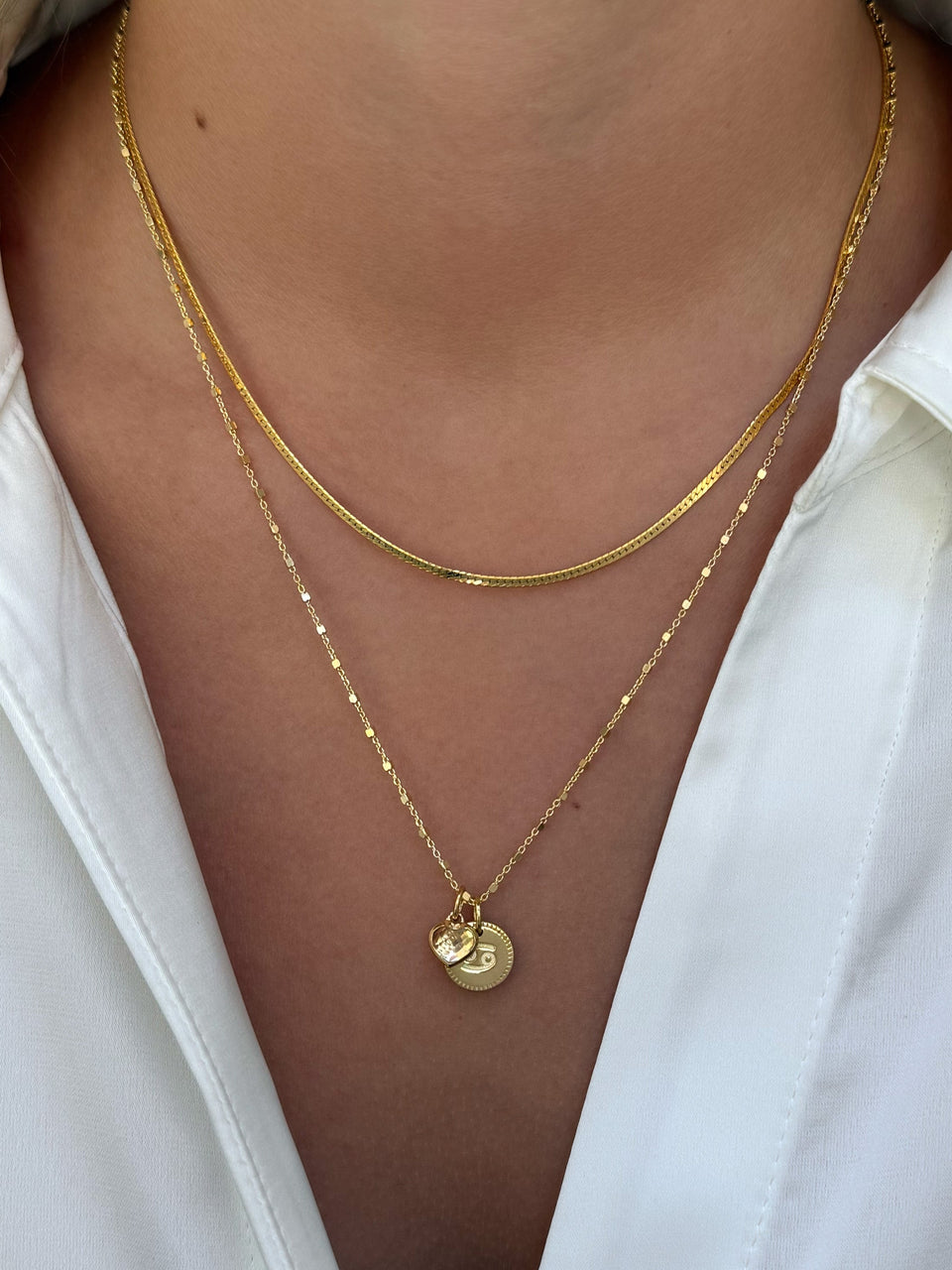 A model wears a layered set of gold necklaces, featuring a flat snake chain and a delicate station chain adorned with a Beloved Zodie Pendant for Cancer and a small Beloved Heart Pendant.