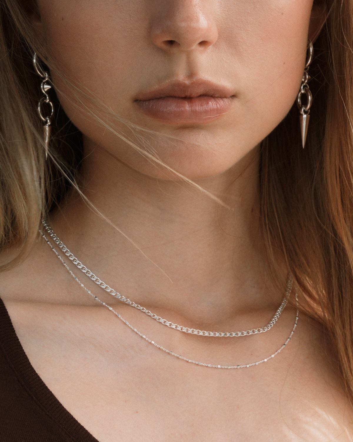 A close-up shot of a model wearing the Sheer Love Necklace in silver, featuring a unique patterned chain, layered with a delicate silver station necklace. She also wears silver drop earrings with a spike design.