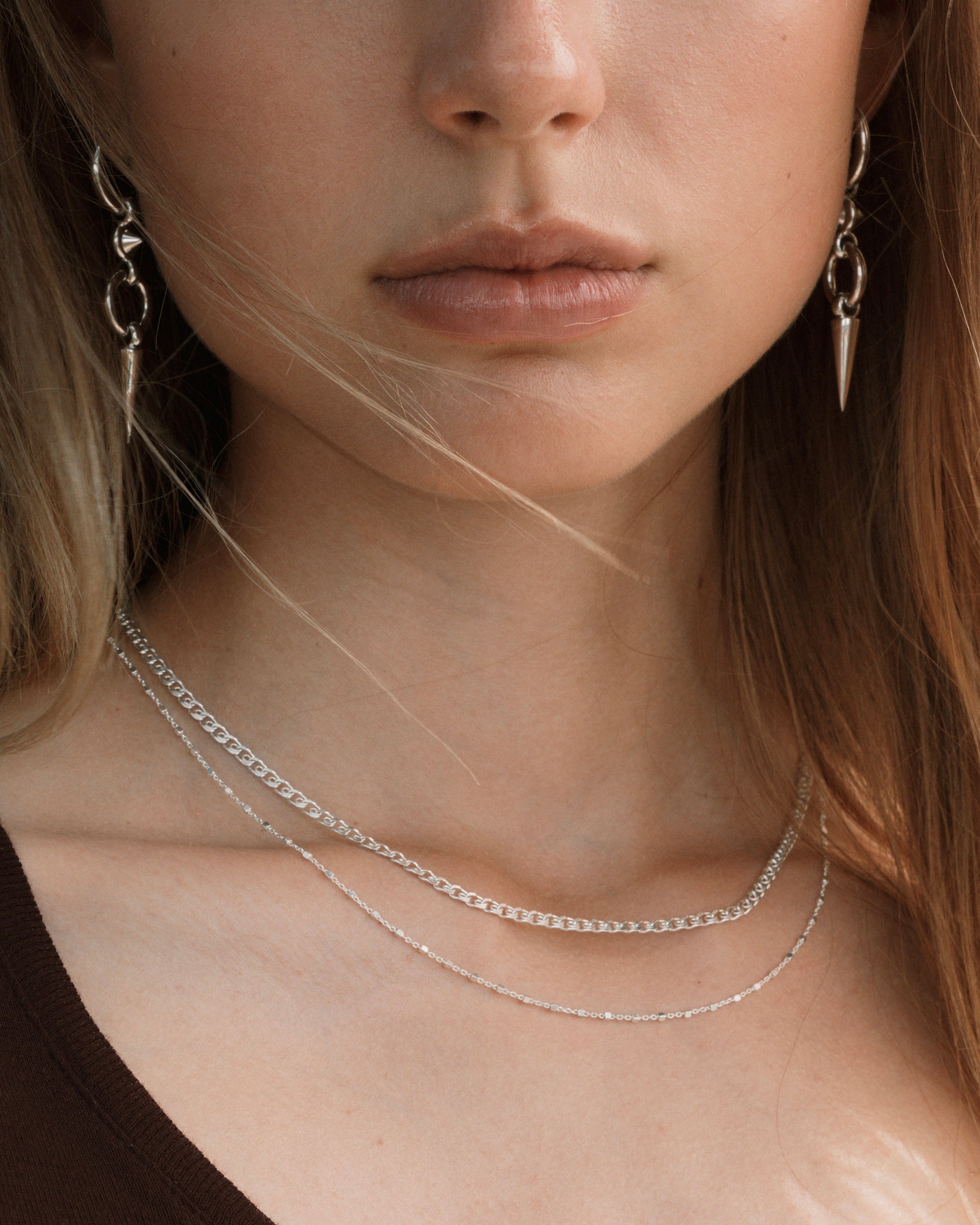 A close-up shot of a model wearing the Sheer Love Necklace in silver, featuring a unique patterned chain, layered with a delicate silver station necklace. She also wears silver drop earrings with a spike design.