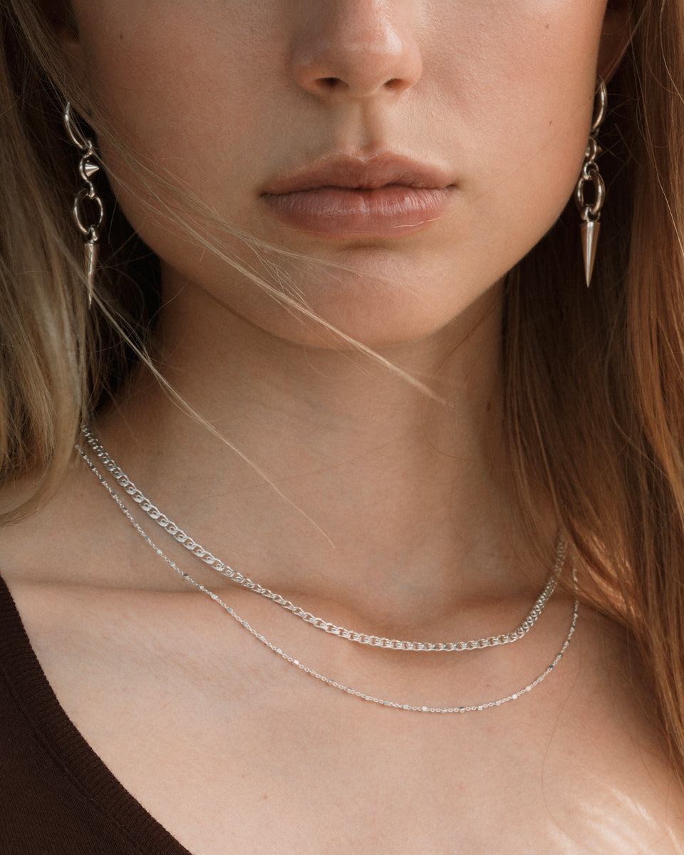 A close-up shot of a model wearing the Sheer Love Necklace in silver, featuring a unique patterned chain, layered with a delicate silver station necklace. She also wears silver drop earrings with a spike design.