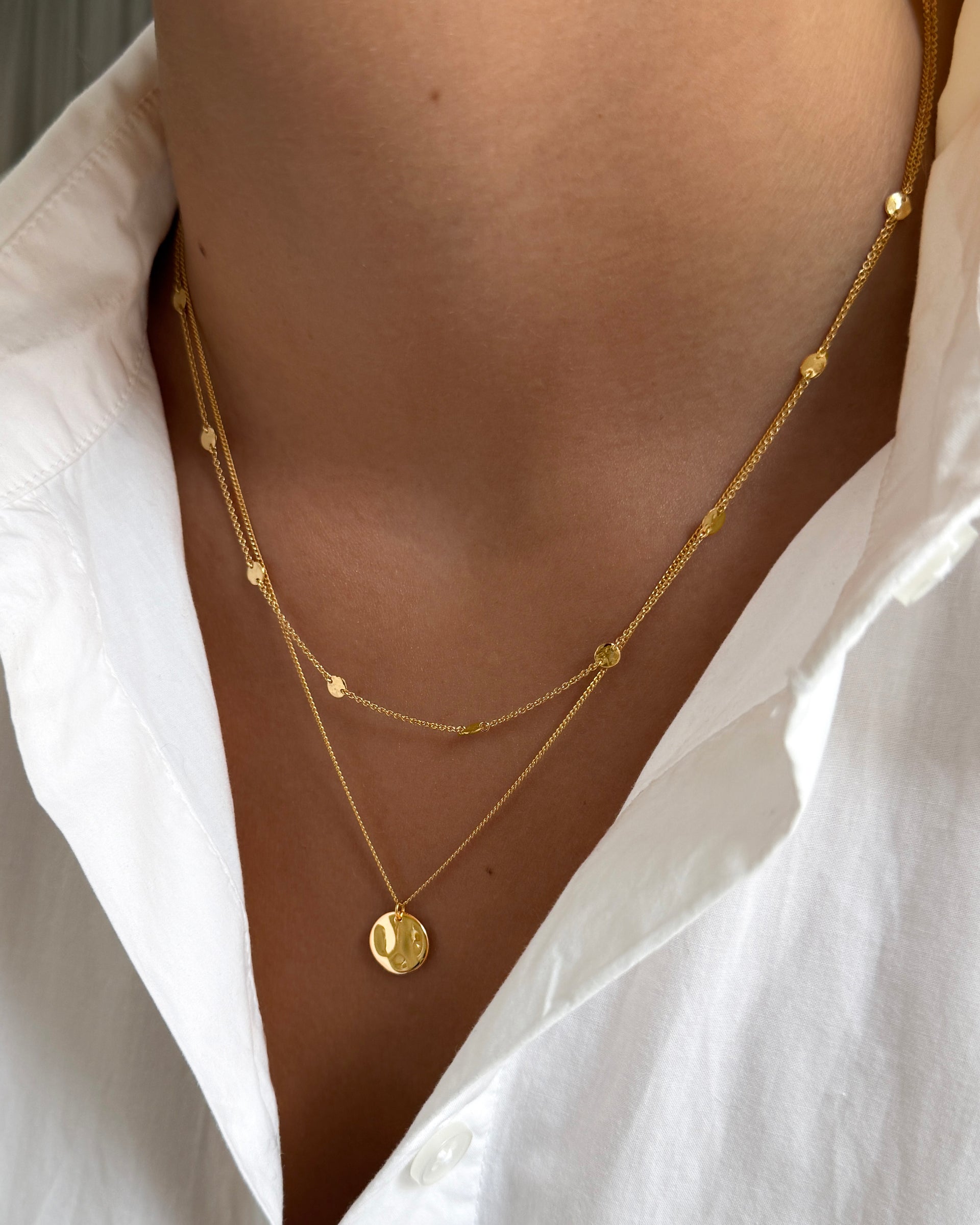 A close-up of a model wearing the Solea Necklace Gold, which has a delicate chain adorned with small hammered gold circles, layered with another gold necklace featuring a round hammered pendant.