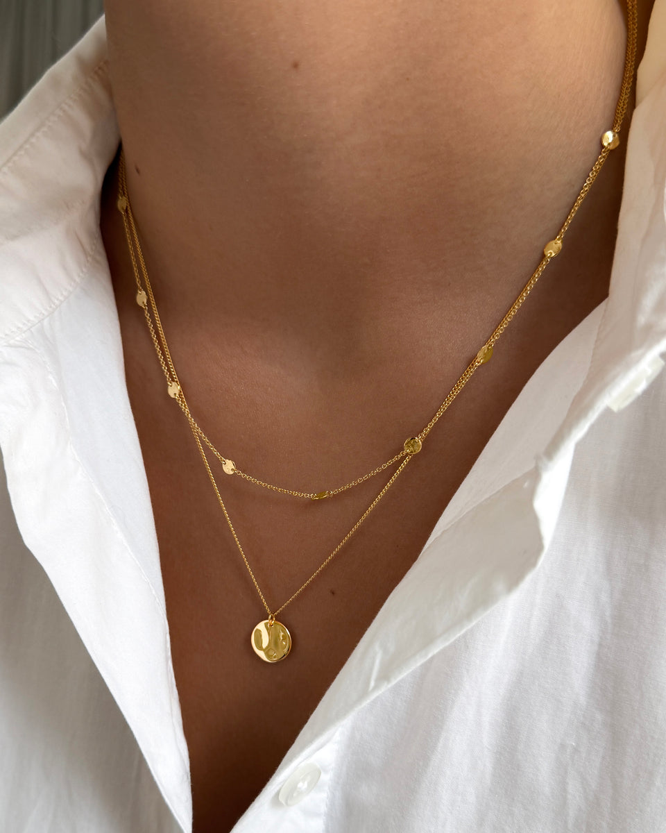 A close-up of a model wearing the Solea Necklace Gold, which has a delicate chain adorned with small hammered gold circles, layered with another gold necklace featuring a round hammered pendant.