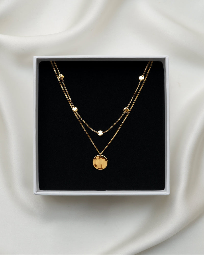 A close-up of a jewelry box with the Solea Necklace Gold, which has a delicate chain adorned with small hammered gold circles, layered with another gold necklace featuring a round hammered pendant.