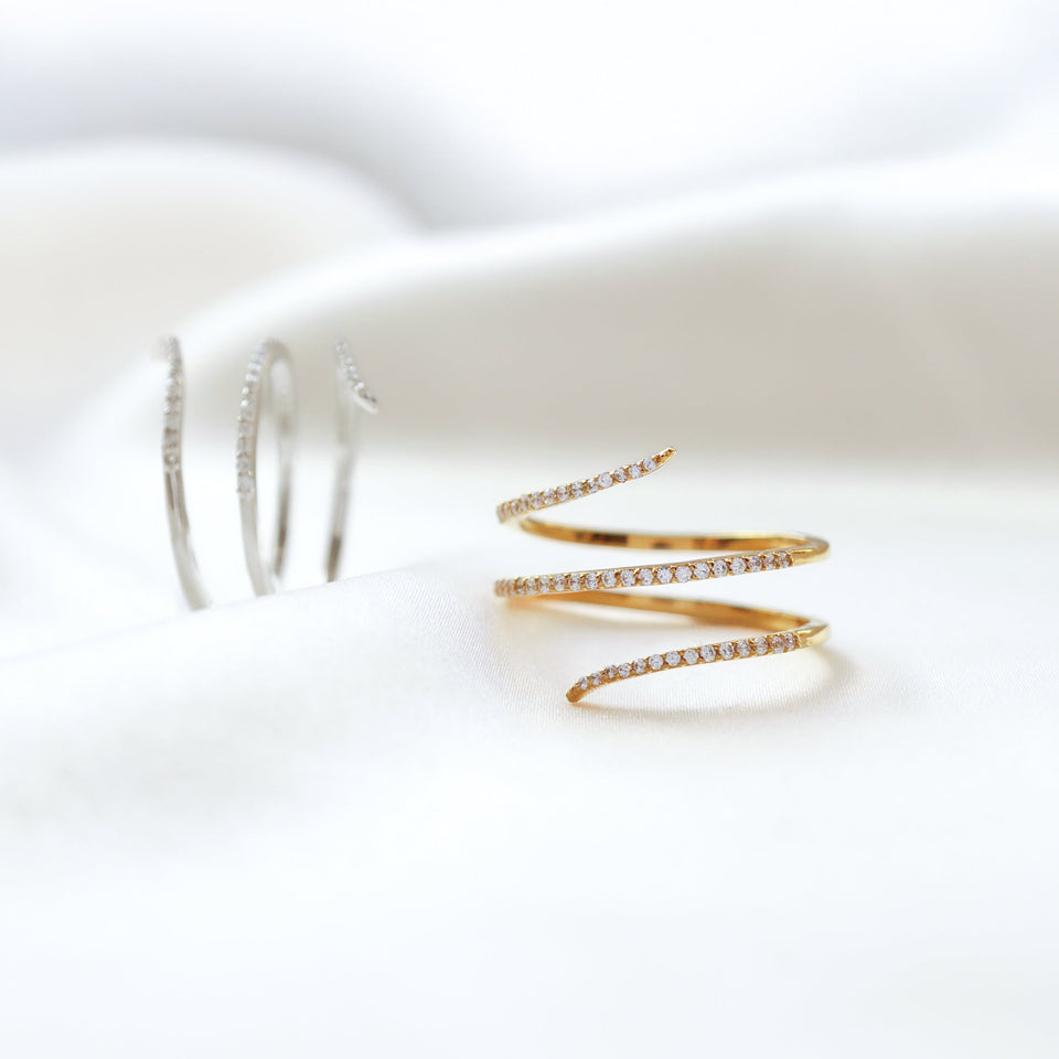 A product shot of the Sparkle Wrap Ring, with the gold version in focus in the foreground and the silver version in the background, both displaying a delicate spiral design set with cubic zirconia.