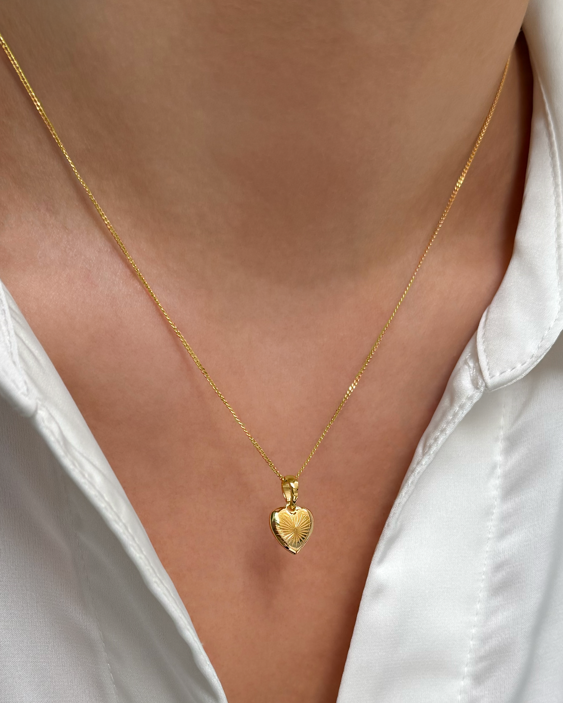 A close-up of a model wearing the Sunburst Heart Necklace in gold, featuring a heart-shaped pendant with a sunburst pattern, styled with a white collared shirt.