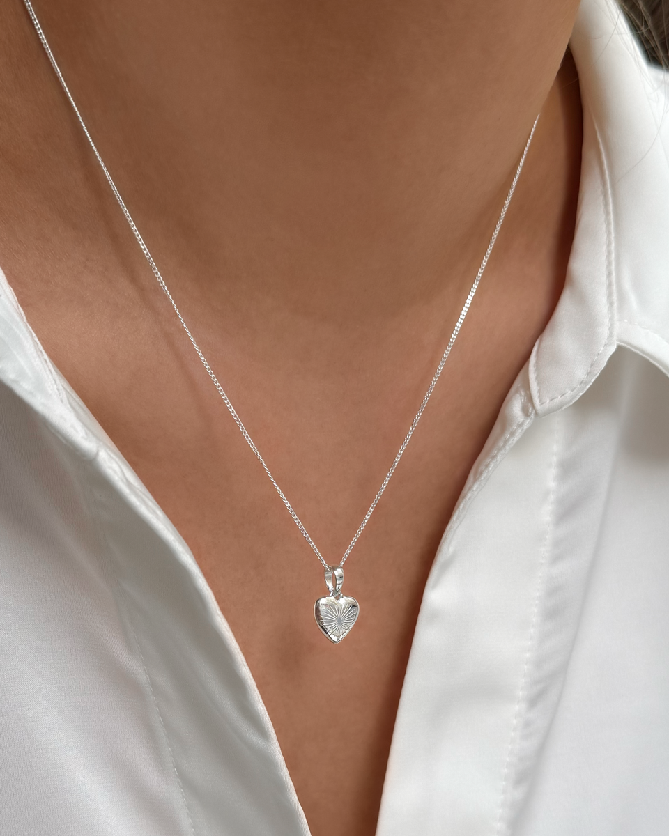 A close-up shot of a model wearing the Sunburst Heart Necklace in silver, featuring a delicate chain and a heart pendant with a sunburst pattern.