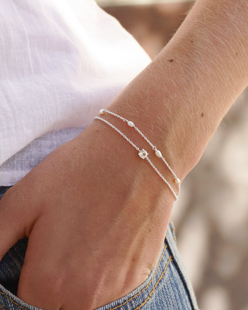 A model wears the Treasure Multi Pearl Bracelet in silver, featuring small freshwater pearls, styled in a layered look with another delicate silver chain bracelet with a single clear stone.