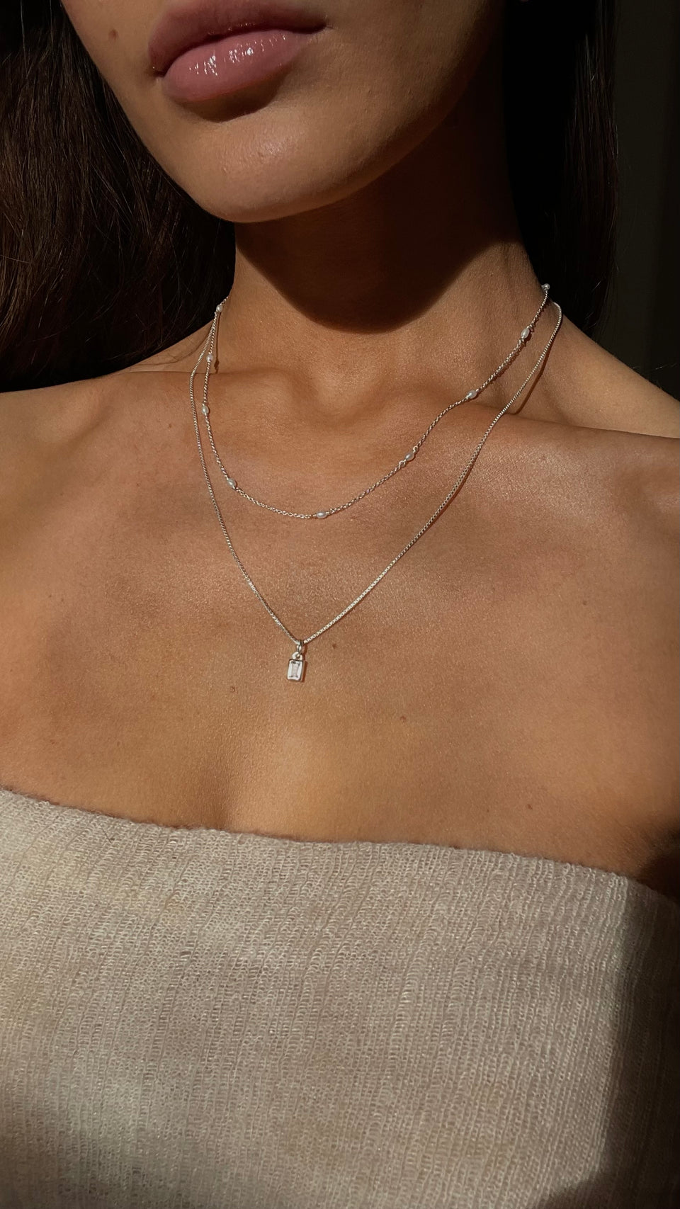 A close-up on a model's neck showing two layered silver necklaces: the Beloved Squared Pendant with a crystal on a fine chain, and the shorter Treasure Multi Pearl Necklace.