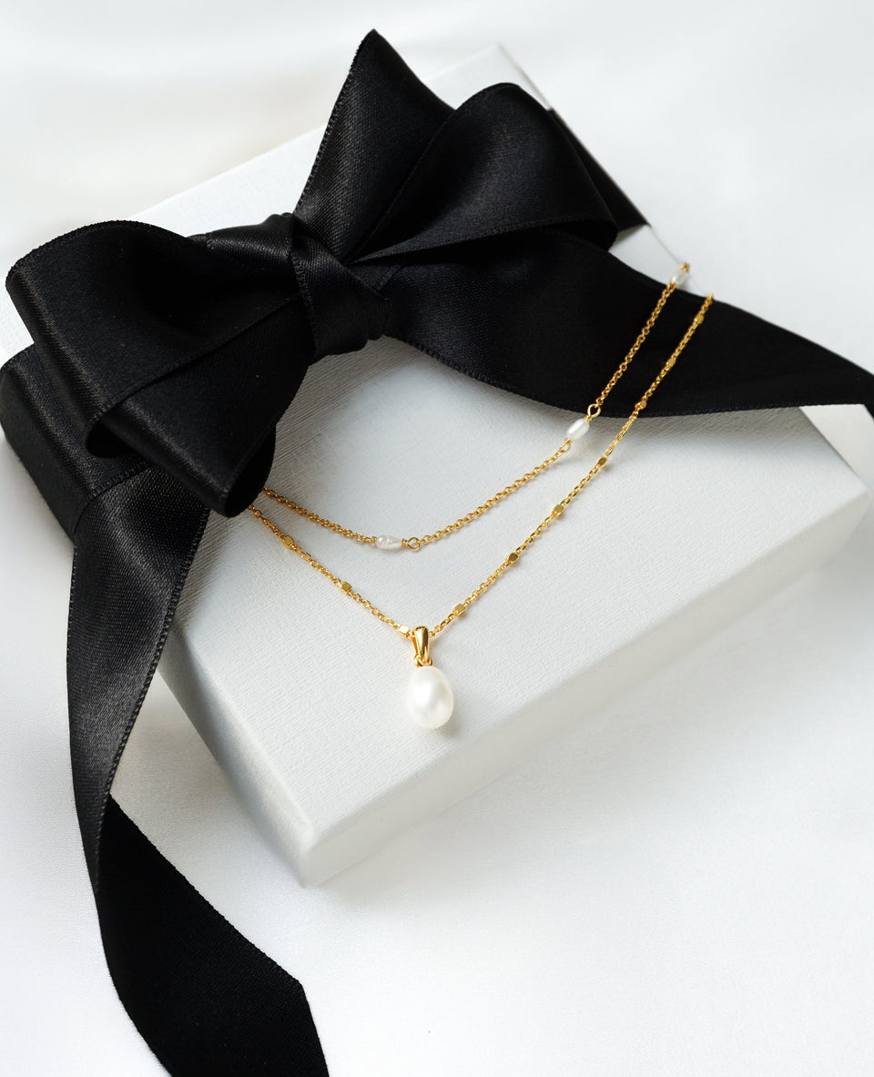 The Treasure Necklace Set Gold displayed on a white Syster P gift box with a large black satin bow. The set features two layered gold necklaces: one with a single teardrop pearl pendant and a shorter one with multiple small pearls.