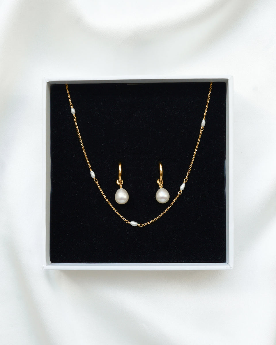 A product shot of the Treasure Necklace & Earrings Set in gold, displayed in its Syster P gift box. The set includes a delicate gold chain necklace with small freshwater pearls and a pair of matching hoop earrings with larger dangling pearl charms.