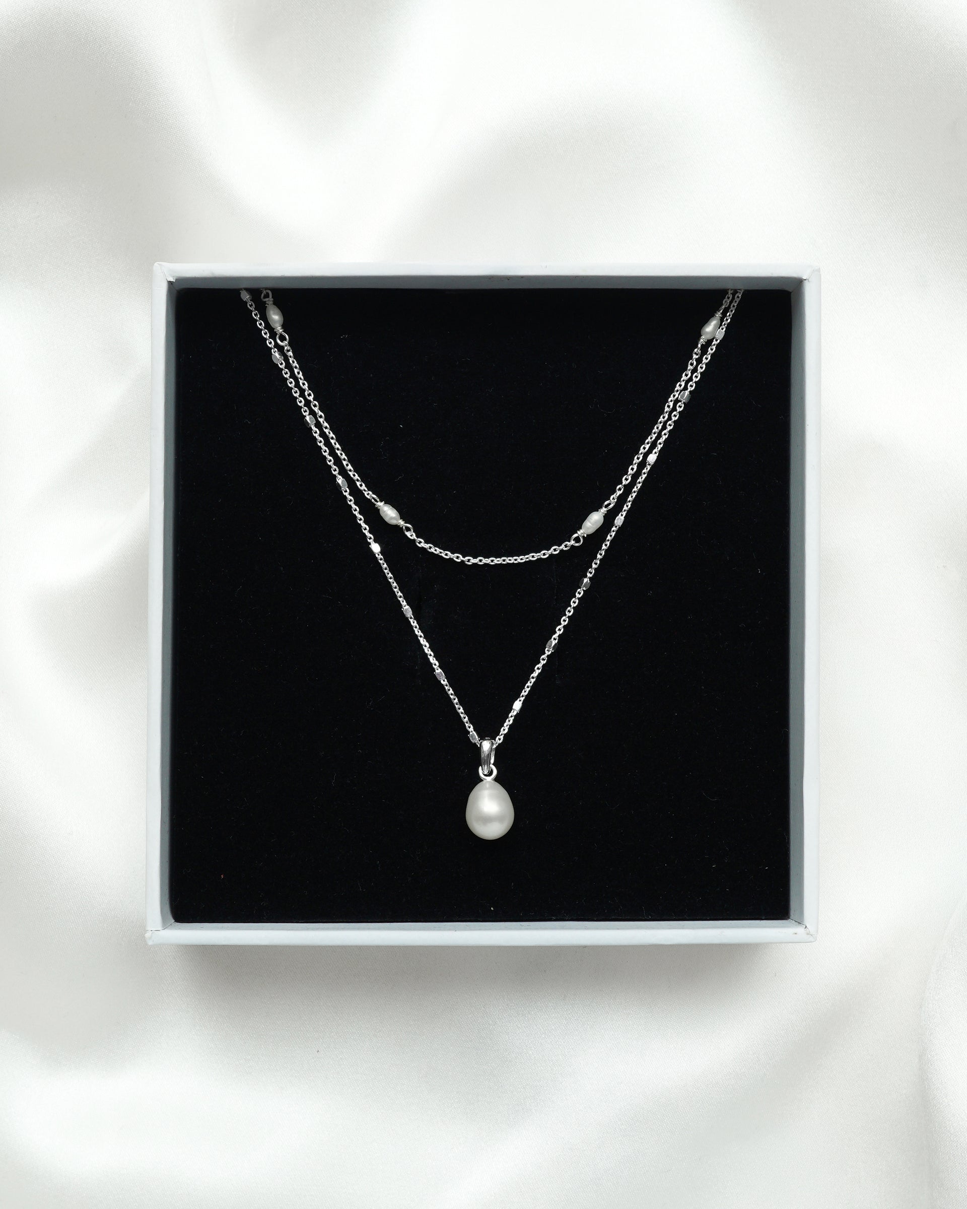 The Treasure Necklace Set Silver is displayed in its white Syster P gift box, showcasing the two layered necklaces: one with a single teardrop freshwater pearl and another with multiple small pearls along its silver chain.