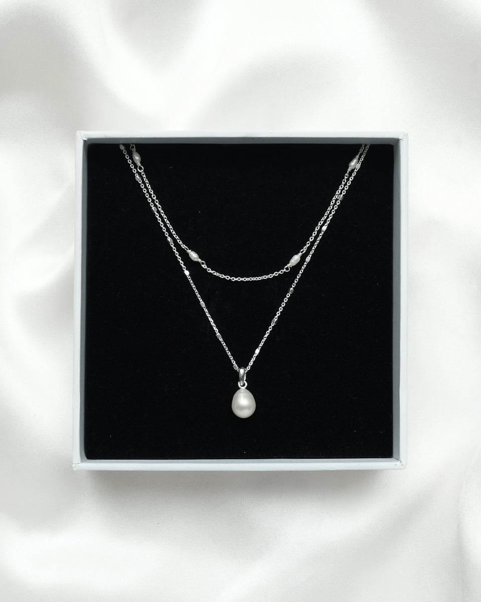 The Treasure Necklace Set Silver is displayed in its white Syster P gift box, showcasing the two layered necklaces: one with a single teardrop freshwater pearl and another with multiple small pearls along its silver chain.
