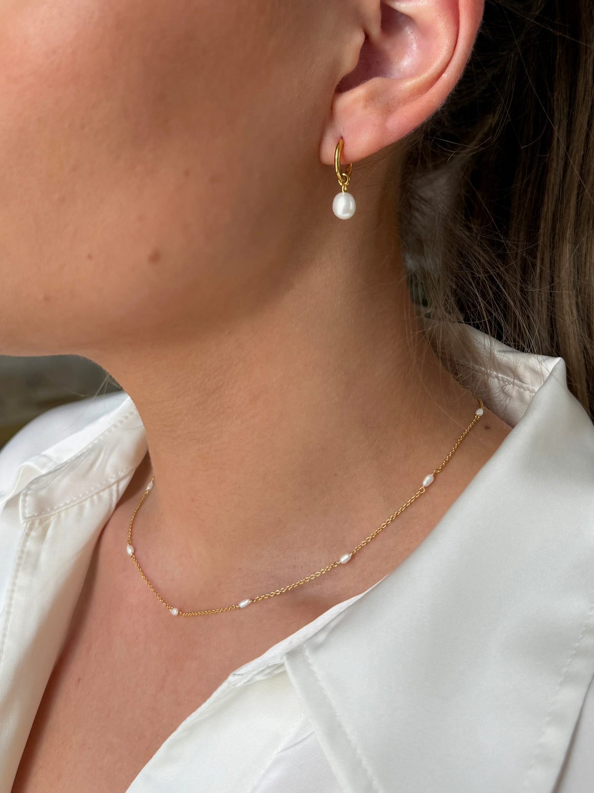 A close-up shot of a model wearing the Treasure Necklace & Earrings Set in gold, featuring a delicate chain necklace with small freshwater pearls and a matching hoop earring with a dangling pearl.