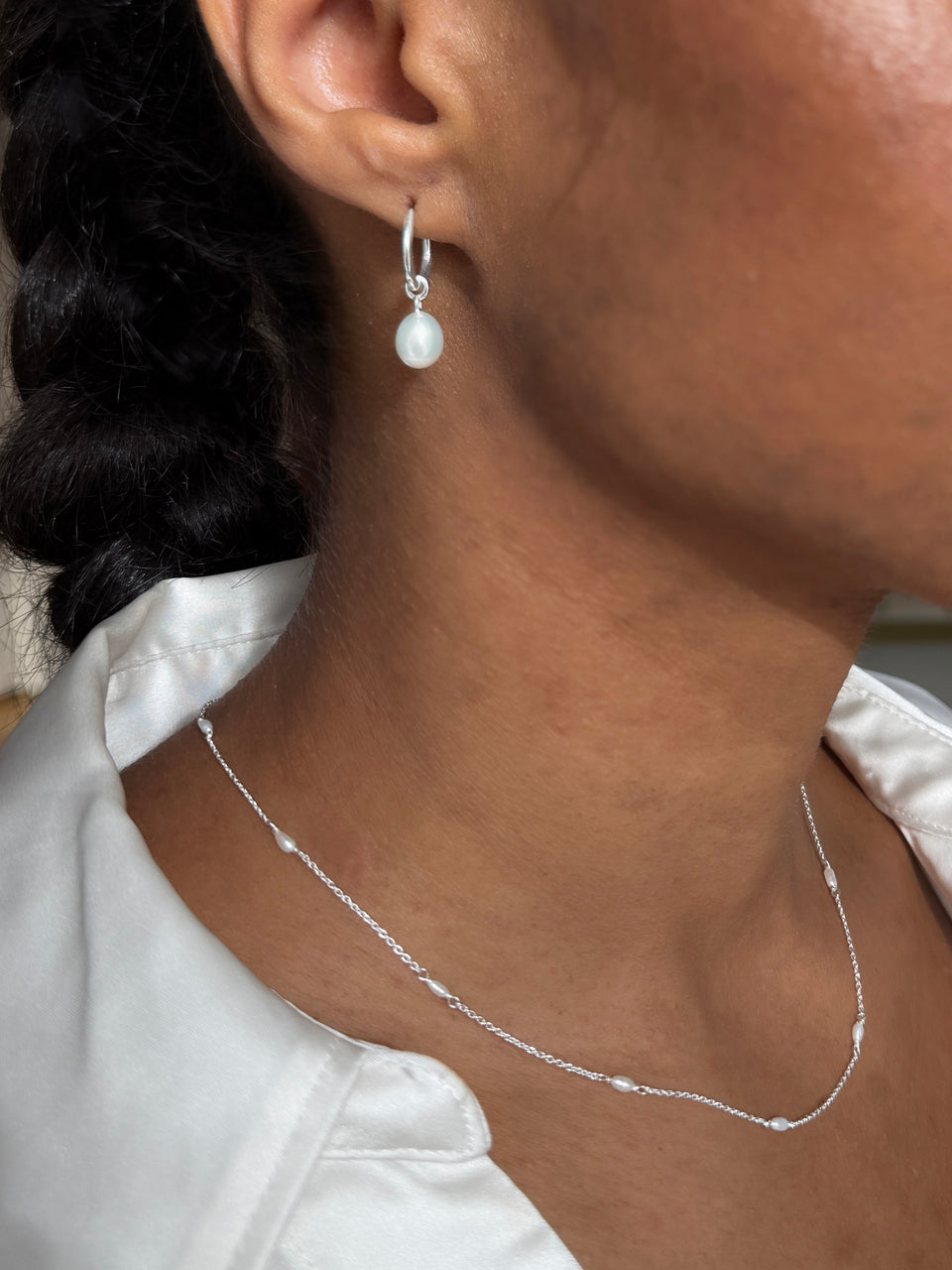 A close-up of a model wearing the Treasure Necklace & Earrings Set in silver, featuring a fine chain necklace with small pearls and a silver hoop earring with a dangling pearl.