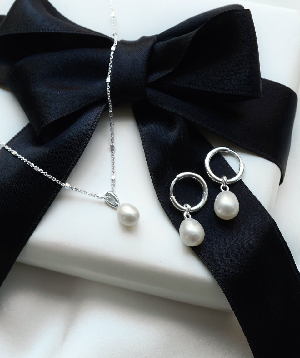 A product shot of the Treasure Necklace & Earrings Set in silver, presented on a white gift box wrapped with a black ribbon. The set includes a delicate silver necklace and a pair of hoop earrings, both featuring a single freshwater pearl.