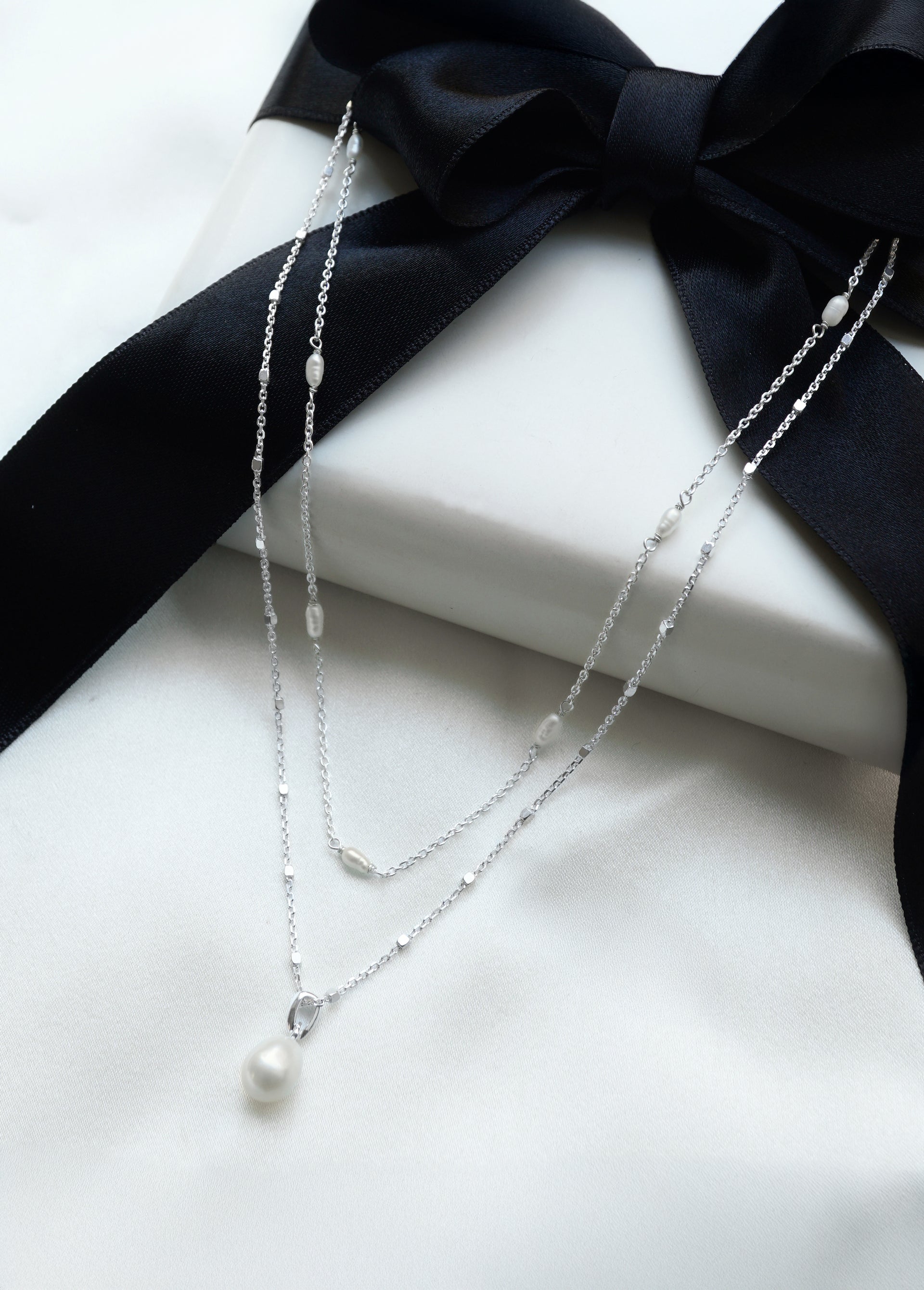 The Treasure Necklace Set in silver, featuring a single pearl pendant necklace layered with a multi-pearl necklace, presented on a white gift box with a black ribbon.
