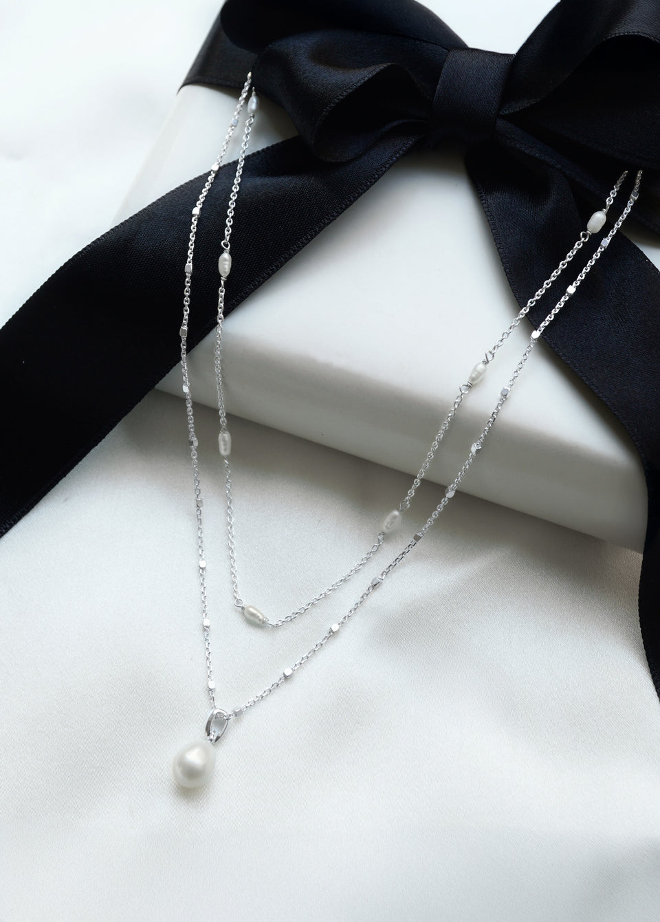 The Treasure Necklace Set in silver, featuring a single pearl pendant necklace layered with a multi-pearl necklace, presented on a white gift box with a black ribbon.