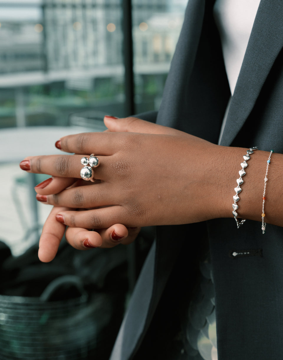 A model wears the Universe Ring Silver, a bold ring with three silver spheres. She styles it with the Darling Bracelet, featuring linked silver hearts, and the Santa Monica Bracelet, a delicate silver chain with colorful beads.